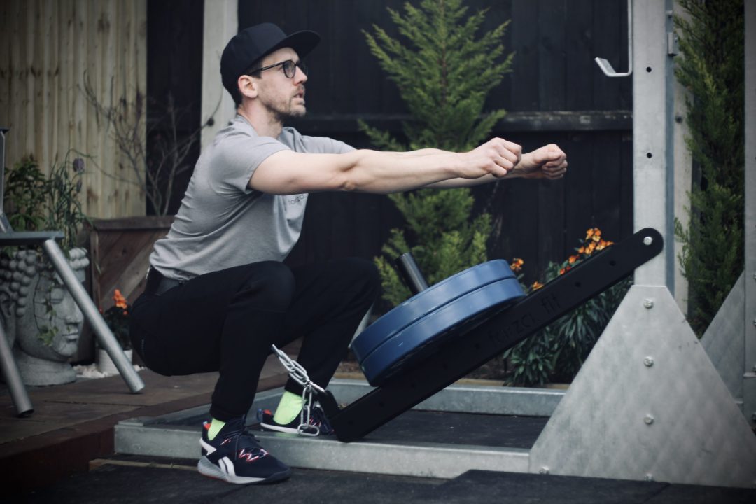 man performing a belt squat with the forte fitness belt squat attachment with 2 blue 20kg plates, on an outdoor rig surrounded by trees