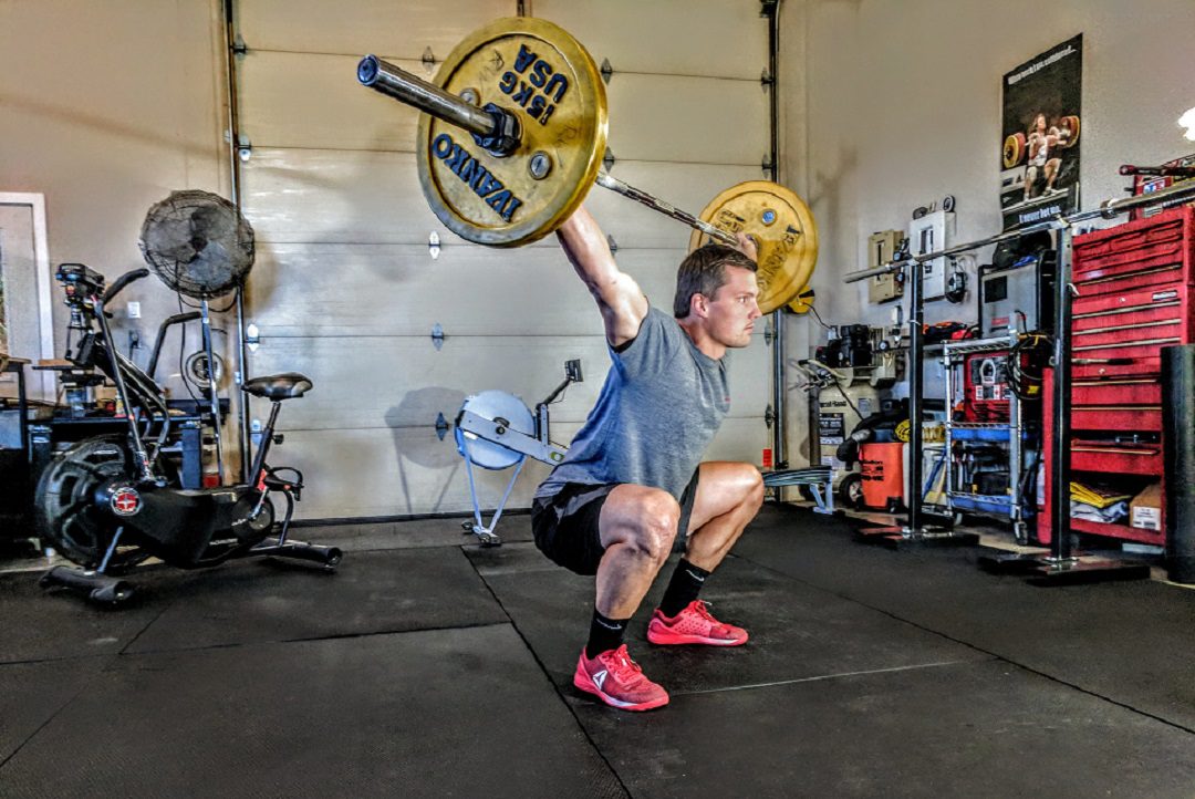 A man squatting in a gym holding weights on a bar above his head.